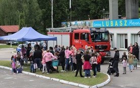 W&oacute;z strażacki wok&oacute;ł kt&oacute;rego stoi duża grupa ludz i z zaciekawienie ogląda go. Obok wozu stoi rozstawiony namiot piknikowy. Jest wiosenny dzień. 